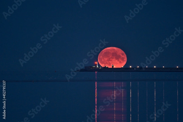 Fototapeta Beautiful view of rare summer full blue sturgeon Moon rising over Irish sky between Dun Laoghaire lighthouses on August 23, 2021 seen from Sandymount Beach, Dublin, Ireland. Huge Moon