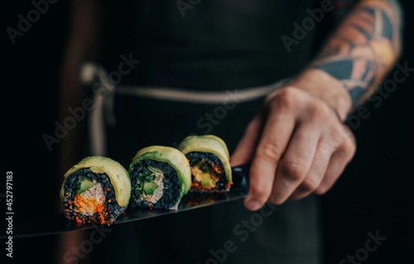 Obraz Unrecognizable man chef with a tattoo on his arm holds sushi rolls on a knife.