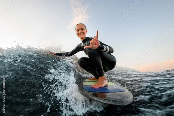 Fototapeta smiling woman sits on wakesurf board and rides the wave and touches the waves with one hand