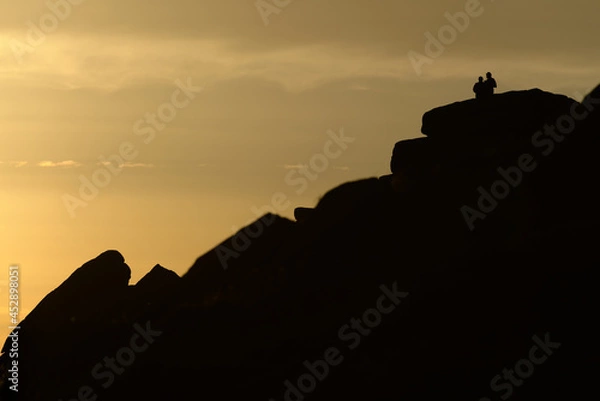 Fototapeta A couple stand silhouetted against a sunset atop stanage edge