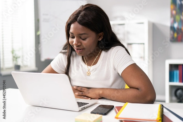 Fototapeta Modest, pretty, dark-skinned girl sits in room at desk, spends afternoon chatting with friends, surfing internet, next to phone, notebooks, women smiles softly at laptop screen, wainting for messages