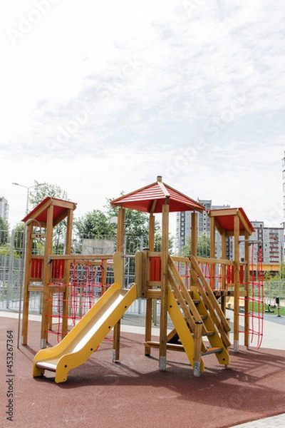 Fototapeta An outdoor colorful playground next to the house. A summer day. Children's playground with rubber floor covering. A wooden house with a slide and ropes.