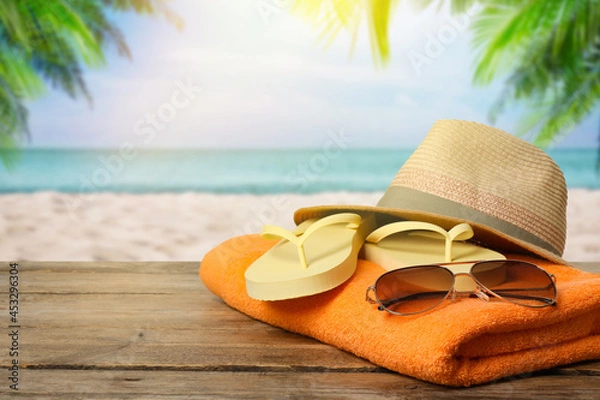 Fototapeta Beach towel, flip flops, hat and sunglasses on wooden surface near seashore. Space for text