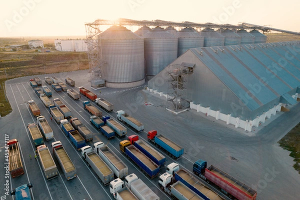 Obraz Grain terminals of modern sea commercial port. Silos for storing grain in rays of setting sun. Many trucks are waiting in line for unloading in port harbor, top view from quadcopter. Logistics, trade