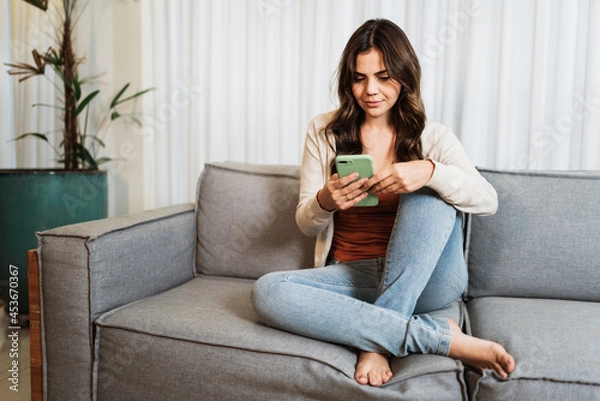 Fototapeta Happy Latin lady relax at home alone sit on room pose share good news at social media via cellphone. Smiling woman enjoy weekend order goods food online in phone app.