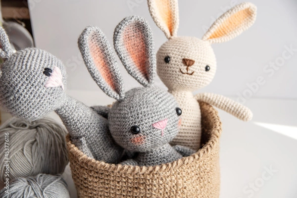 Fototapeta Three handmade bunnies sit in a jute basket on a white table, knitting yarn side by side.
