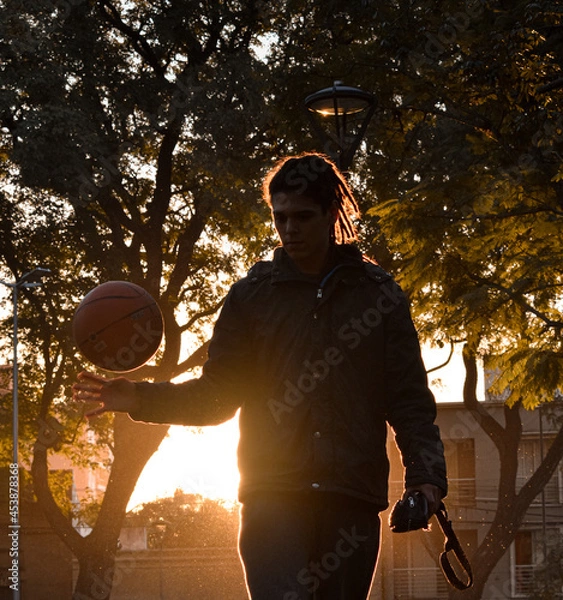 Fototapeta Chico con rastas jugando al baloncesto en un parque durante el atardecer