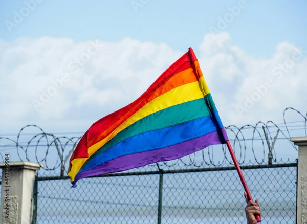 Fototapeta An LGBTQ + flag fluttering in the breeze with a barbed wire fence in the background and a blue sky with clouds.
