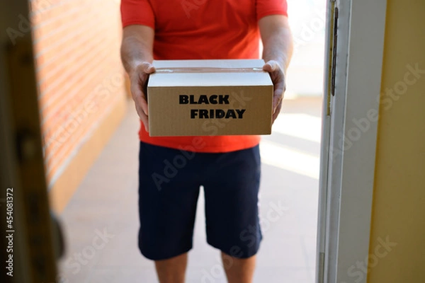 Fototapeta Unrecognizable man delivering a cardboard package marked black friday to the door of a house.