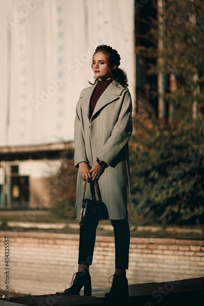 Fototapeta Young fashionable beautiful girl in a long gray coat, black jeans and with a dark red bag. Portrait shooting of a stylish girl. Facial shooting, street style and fashion. Professional model.