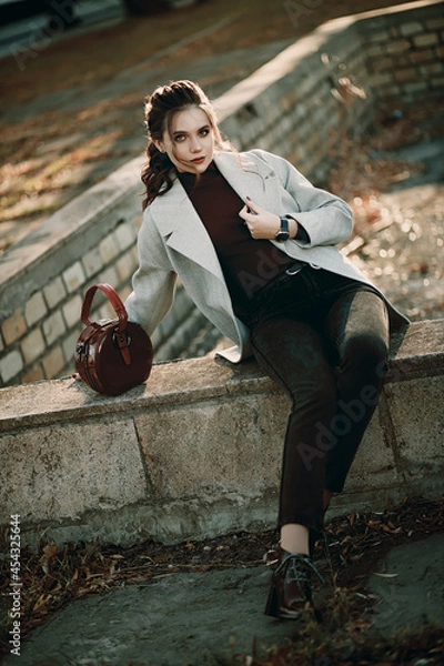 Fototapeta Young fashionable beautiful girl in a long gray coat, black jeans and with a dark red bag. Portrait shooting of a stylish girl. Facial shooting, street style and fashion. Professional model.