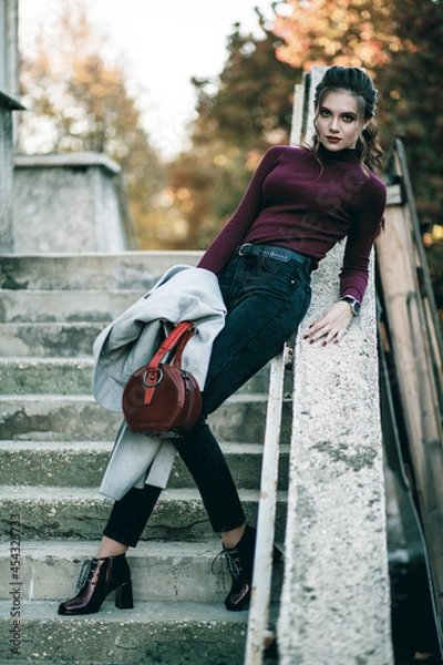 Fototapeta Young fashionable beautiful girl in a long gray coat, black jeans and with a dark red bag. Portrait shooting of a stylish girl. Facial shooting, street style and fashion. Professional model.