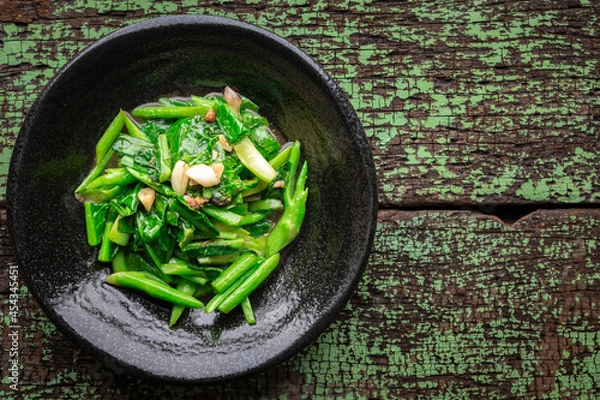Obraz stir fried collards with salted fish and garlic in black ceramic plate on green old wood texture background with copy space for text, top view, collard greens