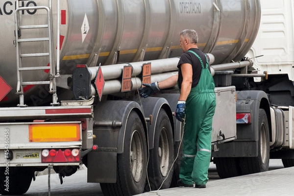 Obraz Belarus, Gomel region - August 21, 2020: A truck driver in overalls grasps the grounding cord or wire to ground the machine. Transportation and delivery of fuel and gasoline. Oil and gas cargo