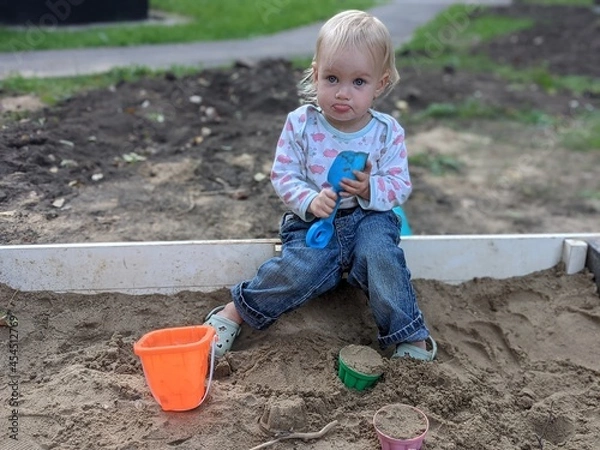 Obraz  a sad child against the background of a poor, unfurnished playground. offended child in the sandbox, with a scoop, mold and bucket