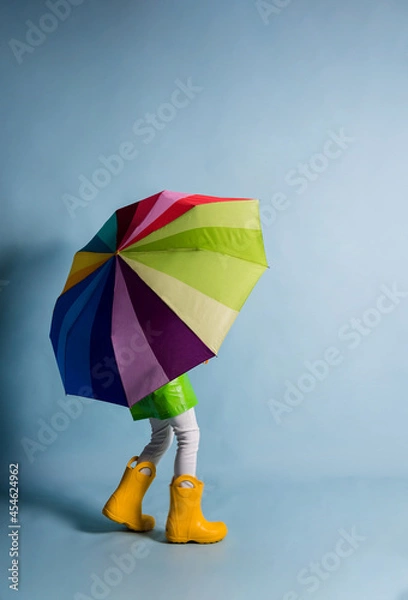 Fototapeta a little boy in a green raincoat and yellow rubber boots covered himself with a multicolored umbrella on a blue background with a place for text