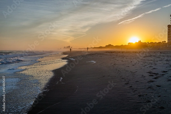 Fototapeta Colorful sunset at Myrtle Beach city by Atlantic ocean with sun behind horizon by condo condominium apartment buildings, people silhouettes walking by crashing waves in resort South Carolina town