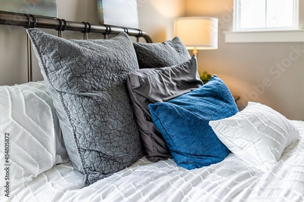 Fototapeta Closeup of decorative pillows cushions in shams and white bedding comforter duvet on bed in bedroom with illuminated lamp on nightstand and bright natural light from window