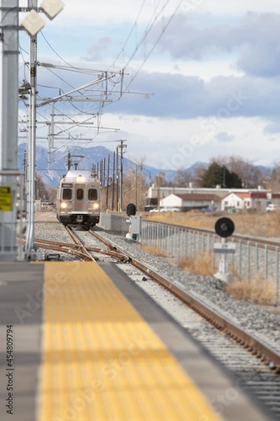 Fototapeta Train arriving at the station. Lights on coming down the tracks. Pedestrian platform in foreground. Rocky Mountains in background.