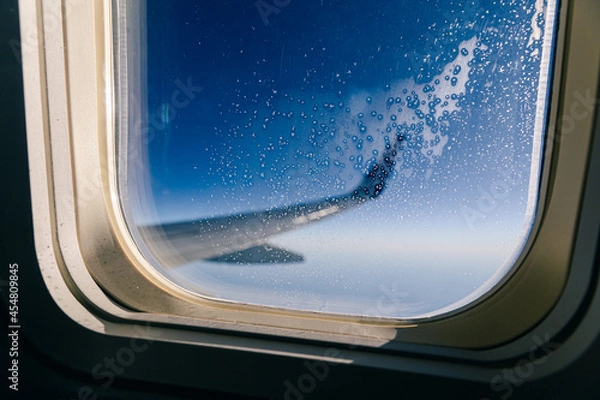 Fototapeta Looking out of airplane porthole. Focus on window frost. Plane wing out of focus in background.