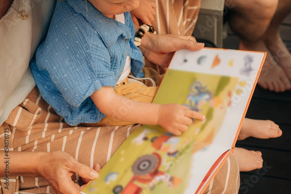 Fototapeta Cute little girl pointing at pictures in book. Toddler sitting on her mother laps and reading book. Family at home, child development concept