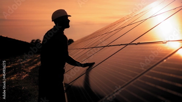 Fototapeta Assistance technical worker in uniform is checking an operation and efficiency performance of photovoltaic solar panels. Unidentified solar power engineer touches solar panels with his hand at sunset