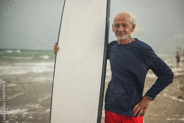Obraz An old man is a surfer with a surfboard. A gray-haired male surfer stands on the beach with a surfboard.