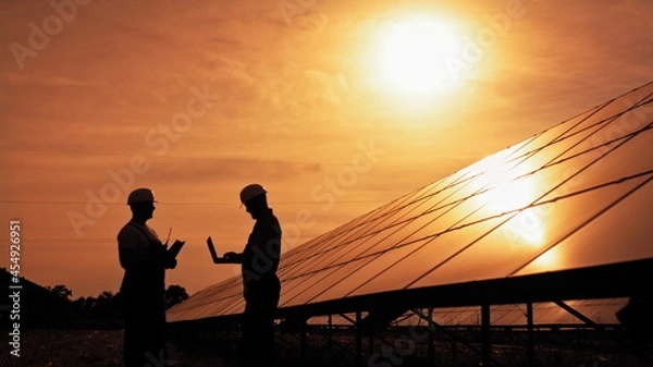 Fototapeta Two unidentified solar power engineers talking to each other at sunset. Couple of workers examining solar batteries and communicating on business. Ecological future. Sustainable energy.