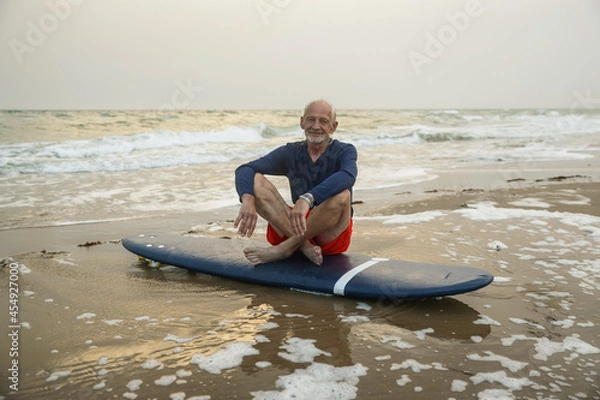 Obraz Handsome adult man walks with a surfboard on the seashore. An old surfer with gray hair sits on a surfboard by the sea and looks to the horizon.