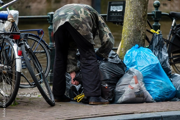 Obraz A man searches for deposit bottles in garbage bags along a canal in Amsterdam, Noord-Holland province, The Netherlands