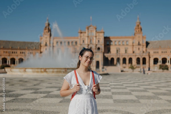 Obraz Young tourist woman with glasses wearing a white dress and red backpack smiles at camera while standing in the Plaza de España of Seville. Blinded by the harsh sun.
