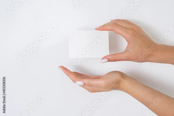 Fototapeta Female hands with a white manicure shows a white empty business card on a light background. Advertising banner for contacts, mockup for beauty salon, business, copy space. Holding information concept