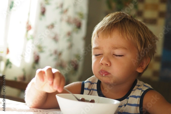 Obraz sleepy boy eating breakfast at the table with his eyes closed early in the morning..