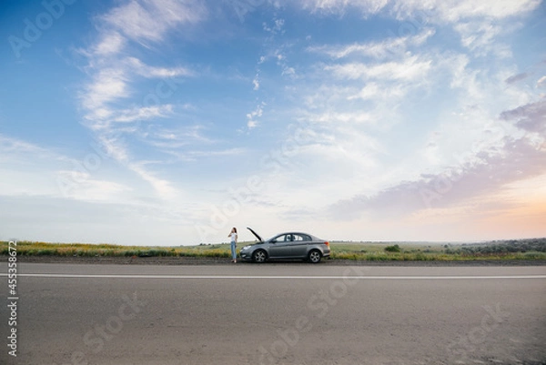 Fototapeta A young girl stands near a broken-down car in the middle of the highway during sunset and tries to call for help on the phone. Breakdown and repair of the car. Waiting for help.