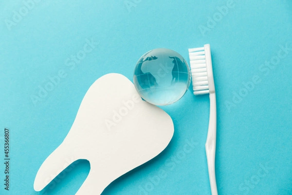 Fototapeta The concept of the International Day of the Dentist. White toothbrush, paper tooth and glass globe on a blue background. Top view. Flat lay. Close-up. Copy space