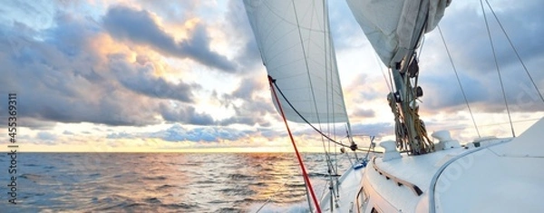 Obraz Yacht sailing in an open sea at sunset. Close-up view of the deck, mast and sails. Clear sky after the rain, dramatic glowing clouds, golden sunlight, waves and water splashes, cyclone. Epic seascape