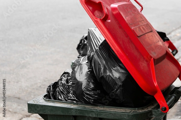 Fototapeta Black plastic bags over flowing modern trash bin with red lid.