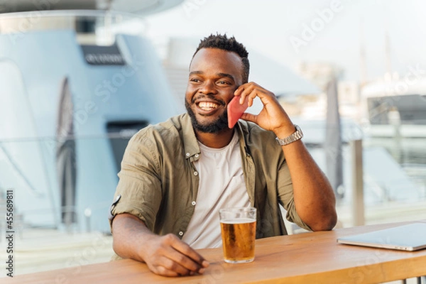 Fototapeta Portrait of a young black man talking to his mobile phone while sitting with a beer and a laptop in a bar.