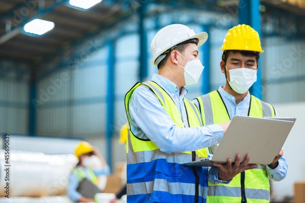 Fototapeta Team engineers and foreman wear a mask, hard hat, and vest. Standing, consult discuss industrial production management through data files with in mask factory. Employees working in the background.