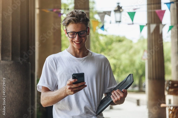 Obraz A man with a smartphone in his hands smiles and writes a message. Business young man wearing glasses with smartphone
