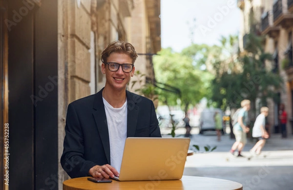 Obraz A young businessman in a suit sits on the terrace of a cafe and works on a laptop. In a big city, a business man sits at a table on the terrace and communicates via a laptop.