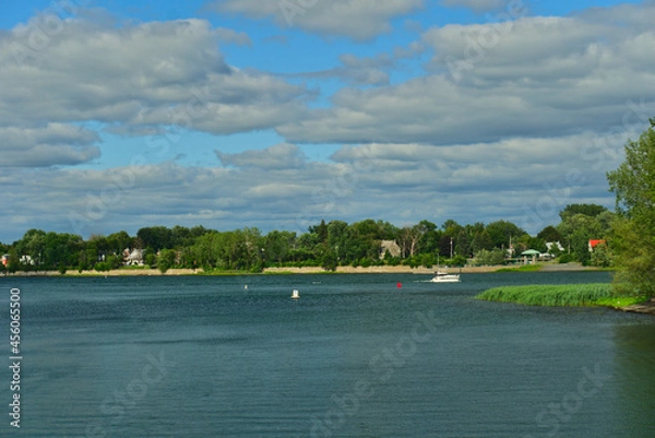 Fototapeta A boat in the river in between the islands of Boucherville national park in Lawrence (Laurent) River, Montreal, QC on a cloudy summer day. The trees and grass on the river banks
