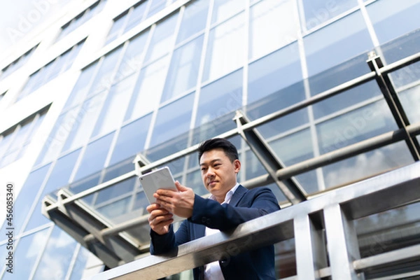 Fototapeta Asian businessman boss reads the news from a tablet computer, standing near his office center on the balcony in a dark color
