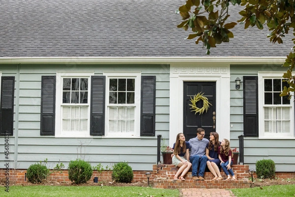 Fototapeta A family with a mother, father, and two daughters sitting outside on the brick steps of a front porch of a small blue cottage house