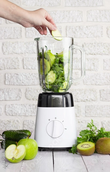 Obraz Blender with green fruits and vegetables on a light background. Preparation of a healthy smoothie. The hand puts a piece of apple in the blender bowl.