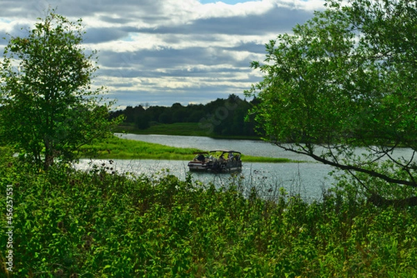 Fototapeta A boat in the river in between the islands of Boucherville national park in Lawrence (Laurent) River, Montreal, QC on a cloudy summer day. The trees and grass on the river banks