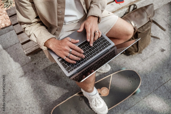 Fototapeta Faceless portrait of young man skater sitting in the street working on laptop with mobile phone with longboard and backpack. Remote working concept. Millennial hipster guy. Hands typing from above
