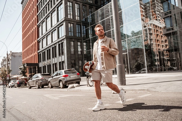 Fototapeta Attractive man skateboarder holding longboard on shoulder and cup of coffee takeaway looks seriously focused at distance while crossing the road on urban building background. Wears stylish outfit.