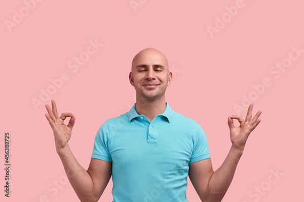 Fototapeta Peaceful pleased bald homosexual man with bristle is practicing yoga, relaxing, makes zen gesture, meditates indoors , closes eyes, gay friendly, wears blue polo shirt, poses over pink background.