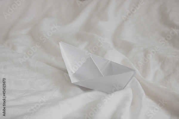 Obraz paper origami boat in bed with waves made with bed sheets, natural white crumpled blanket